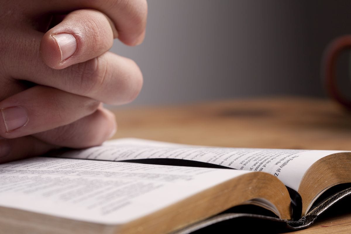 Hands of a man praying over a Bible at his desk.